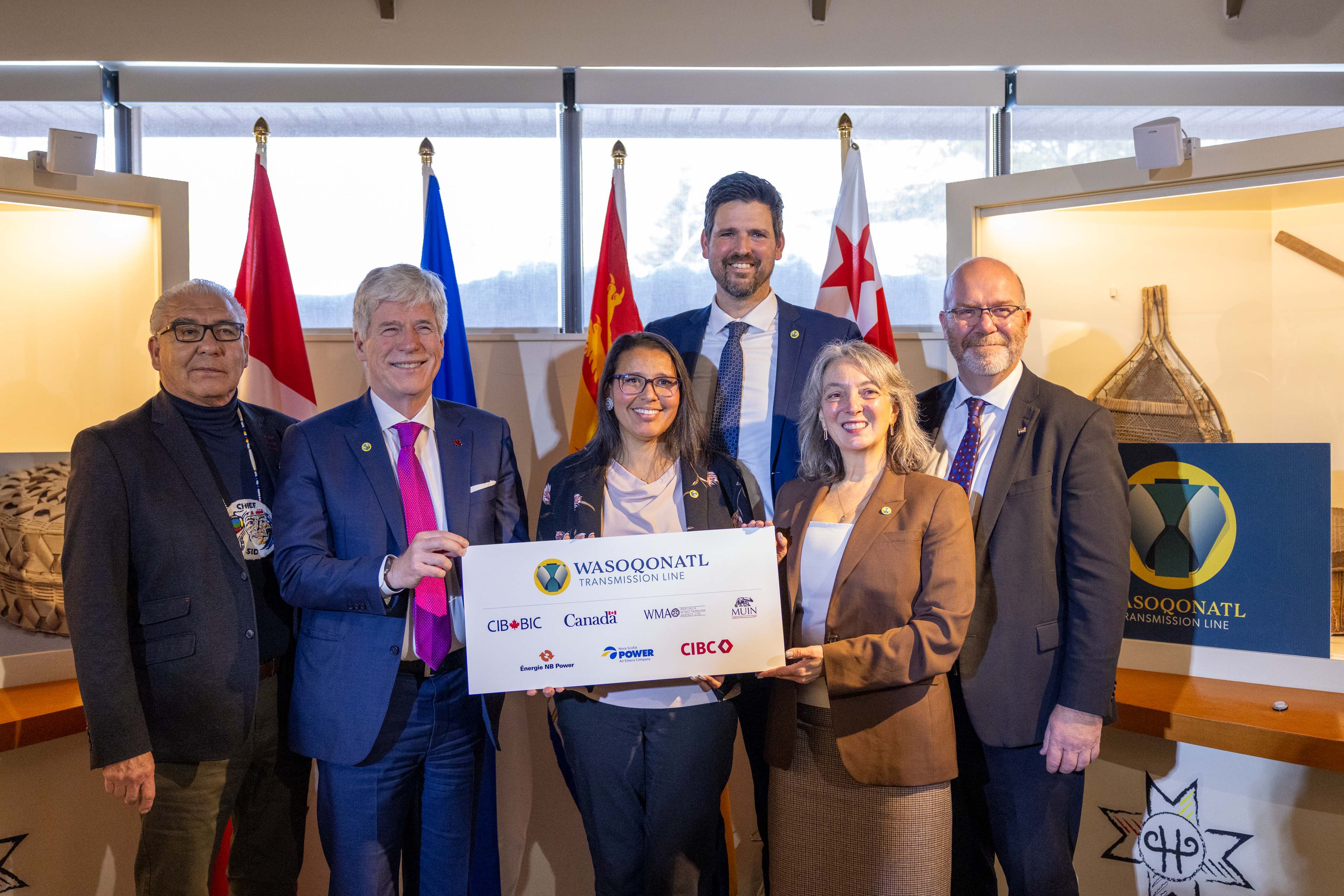 Six officials in formal attire holding a large ceremonial check, standing in front of flags including Canadian and provincial flags in an institutional setting.