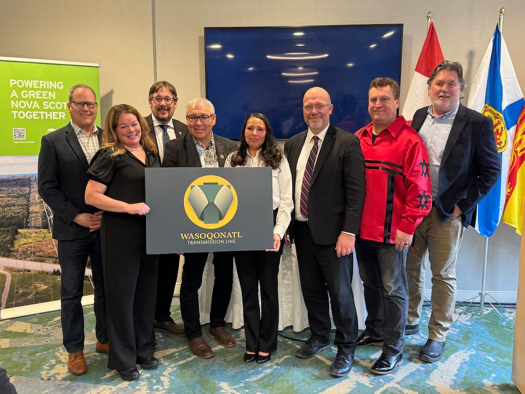 Group of nine professionals in business attire holding a yellow emblem logo at an indoor event, with Canadian and provincial flags visible in the background.